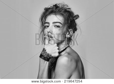 Pretty girl or beautiful woman young cute sexy fashion model with curly hair nose ring and necklace with cuffs poses topless with thumb on lips on grey background