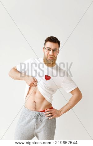 Handsome muscular man raises his white t shirt, demonstrates naked body, poses against white studio background. Positive bearded male model in spectacles shows perfect masculine body. Vertical shot.