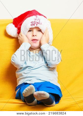 Happy childhood. Full length of blond boy child kid preschooler in red santa hat sitting on sofa with yellow blanket. Christmas at home.