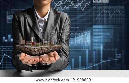 Cropped image of businessman in suit presenting in his hands modern city block with business sketches on background. 3D rendering.