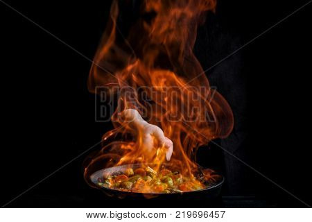 Chef is stirring vegetables in wok with fire. Dark black background.