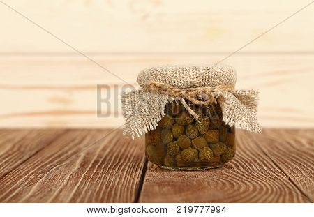 Close up of one small glass jar of pickled capers with canvas top decoration and twine on brown wooden table over white background low angle side view