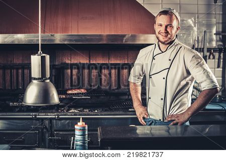 Food concept. Young smiling and handsome chef in white uniform keeps his hands on his belt standing in the interior of modern restaurant kitchen. Preparing traditional beef steak on barbecue oven