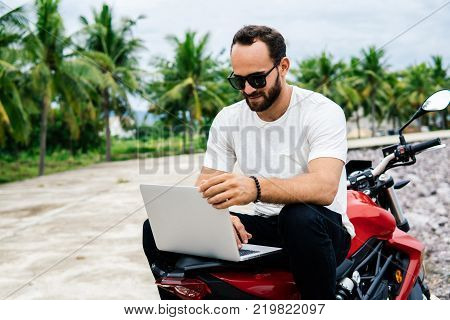 Man working on laptop computer while sitting on his motorcycle on palm background