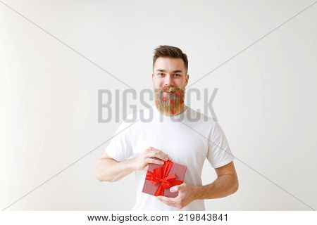 Cheerful bearded male holds wrapped gift box, dressed in casual white t shirt, has beard decorated with paper red hearts, glad to recieve present, celebrates his birthday together with friends.