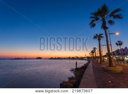 View over the seafront of the city Paphos in Cyrpus. Palm silhouettes, boats in the water and golden sunset over the sea with dramatic cloudy sky.