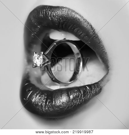 Closeup of female sexy lips with red lipgloss holding round beautiful gold wedding ring as valentine day present with gem in opne mouth with tongue and white teeth square photo