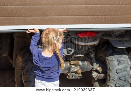 Little blond girl opening garage door where ATV stands. Back view.