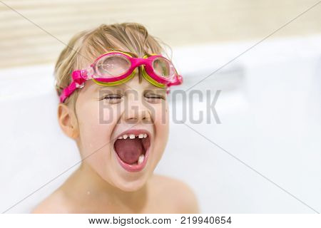 Little cute caucasian girl with blond wet hair wearing pink swimming goggles having fun and peaking out of bath side with tricky sight.
