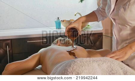Spa therapist pouring hot chocolate on young woman back at luxury beauty salon. Beautiful young woman at skin care salon. Professional chocolate massage.