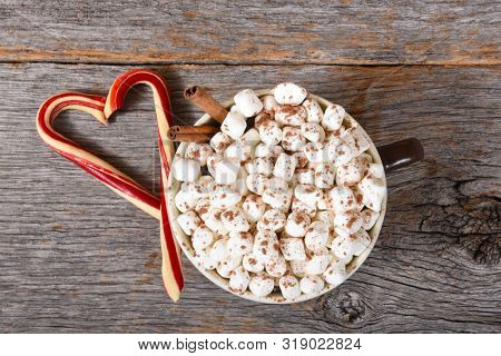High angle shot of a large mug of hot cocoa with marshmallows and two candy canes forming a heart on a rustic wood table.