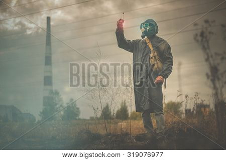 Man In Gas Mask Is Looking A Radioactive Liquid In Vial In His Hand On A Smoking Chimney Background.