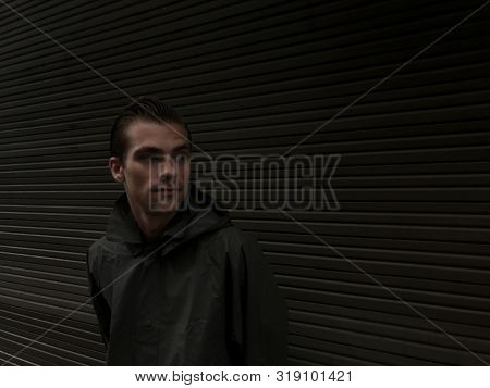 Handsome Young Man In A Raincoat Stands In Front Of A Wall, Garage Doors. Cope Space, Place For Text