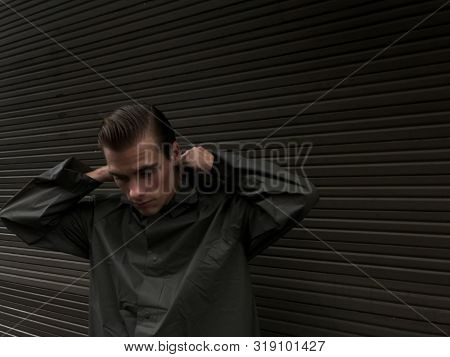 Handsome Young Man In A Raincoat Stands In Front Of A Wall, Garage Doors. Cope Space, Place For Text