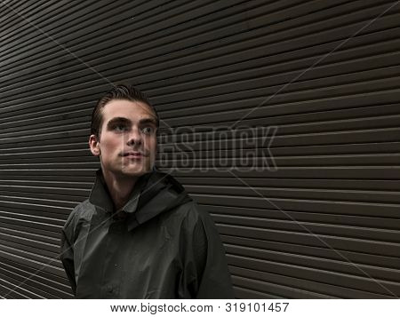 Handsome Young Man In A Raincoat Stands In Front Of A Wall, Garage Doors. Cope Space, Place For Text