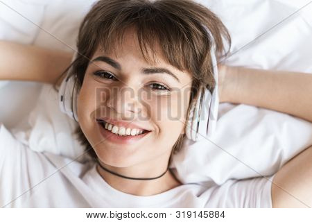 Photo of a happy optimistic positive pleased young beautiful woman lies indoors at home on bed listening music with headphones.