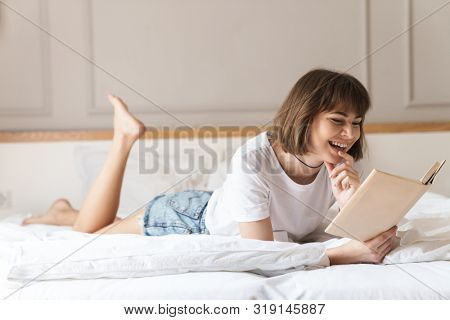 Image of a pretty happy cheerful positive young beautiful woman indoors at home lies on bed reading book.