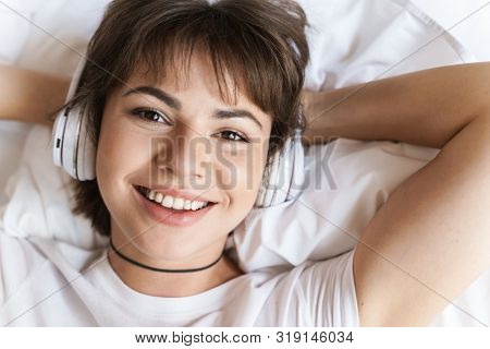 Photo of a cheerful optimistic young beautiful woman lies indoors at home on bed listening music with headphones.