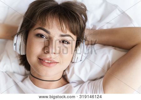 Photo of a cheerful optimistic young beautiful woman lies indoors at home on bed listening music with headphones.