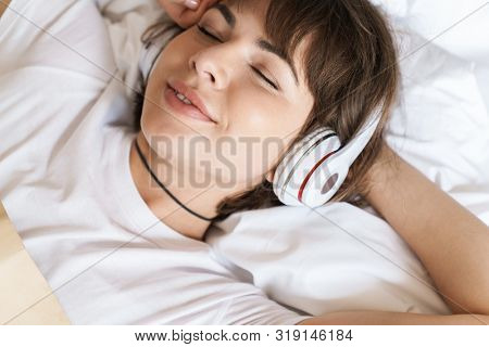 Photo of a pleased relaxing optimistic young beautiful woman lies indoors at home on bed listening music with headphones.