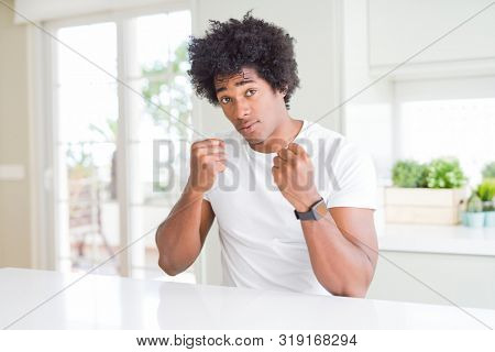 Young african american man wearing casual white t-shirt sitting at home Ready to fight with fist defense gesture, angry and upset face, afraid of problem