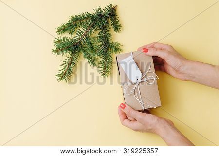 Female Hands, Placing Gift Box Next To Fir Branch Over Yellow Background