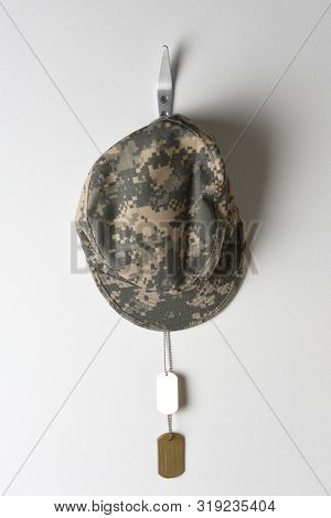 A set of military dog tags and field cap hanging from a hook on a blank wall.