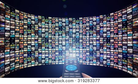 Image of big screen with many multi-colored channels, hands of man with remote control.