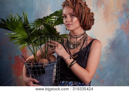 Southern Flavor. A Young Tanned Woman With Afro Braids Smiles Holding A Big Pot With A Tropical Plan