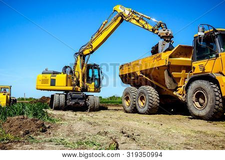 Big Excavator Is Filling A Dumper Truck With Soil At Construction Site, Project In Progress.