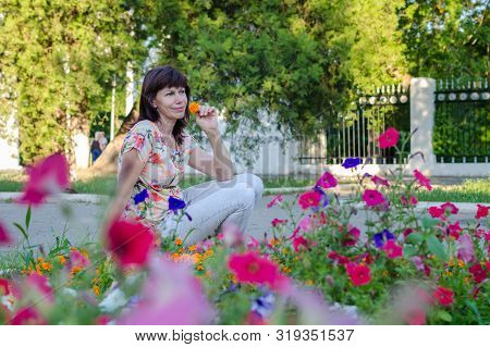 Portrait Of A Beautiful Happy Lonely Elderly Woman Against A Flower Bed. Walks In The Park, Dreams O