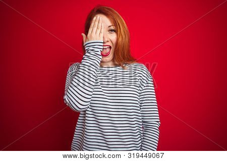 Young redhead woman wearing strapes navy shirt standing over red isolated background covering one eye with hand, confident smile on face and surprise emotion.