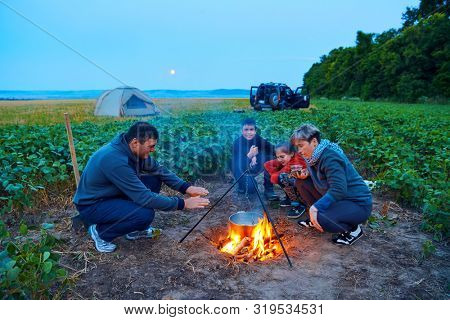 Family traveling and camping, twilight, cooking on the fire. Beautiful nature - field, forest and moon.