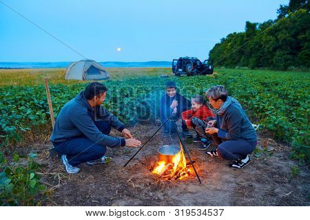 Family traveling and camping, twilight, cooking on the fire. Beautiful nature - field, forest and moon.