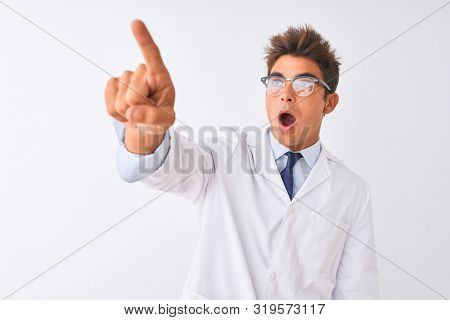 Young handsome sciencist man wearing glasses and coat over isolated white background Pointing with finger surprised ahead, open mouth amazed expression, something on the front