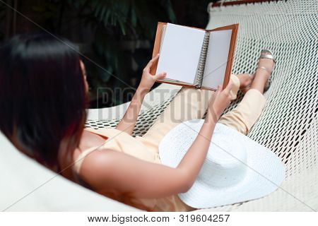 Young And Cute Asian Girl Lying In A Hammock And Reading A Book During Holiday