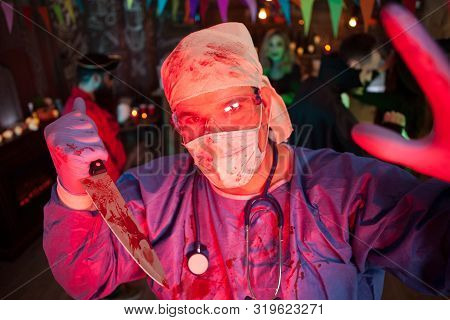 Creepy Doctor With His Covered Holing A Kinfe With Blood And Looking Into The Camera At Halloween Pa