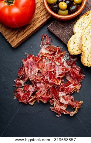 Appetizing Slices Iberian Ham In The Foreground. Olive Oil, Bread, Fresh Tomato, Olives. Black Backg