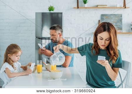 Woman With Smartphone Overfilling Bowl With Milk While Sitting At Kitchen Table Near Husband And Dau