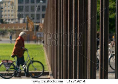 Elder Berliner Cyclist Woman Dressed In A Brown Coat Near A Grass Field Looking At The Metal Bars Of