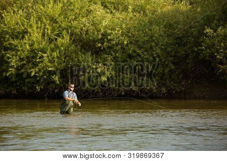The Fisherman Stands At Waist-deep In Water. A Man In A Special Waterproof Suit Throws A Fishing Rod