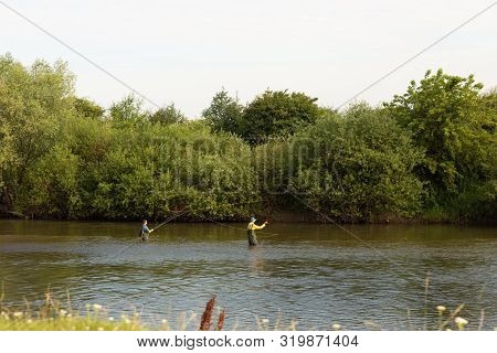 Two Fisherman Stands At Waist-deep In Water. Man In A Special Waterproof Suit Throws A Fishing Rod
