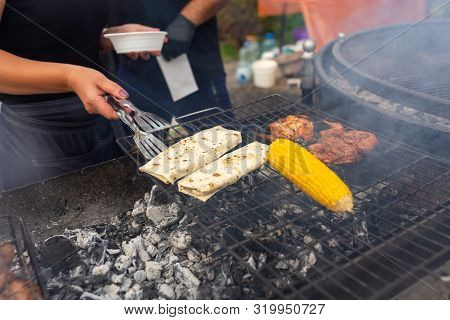 The Chef Turns Over With A Spatula Two Pita Breads Which Are Fried On The Grill Next To Corn. Food A