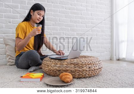 Asian Female Sit On Floor Using Laptop On Wicker Stand And Drink Orange Juice In Living Room At Home