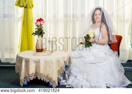 A Joyful Jewish Bride With Her Face Covered With A Veil With A Bouquet Of White Roses Sits In A Syna