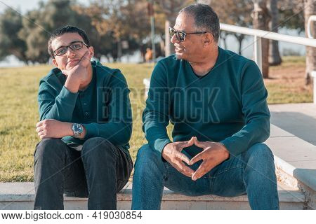 Teenager Son And Senior Father Sitting On Stairs Outdoors At Home And Talking.
