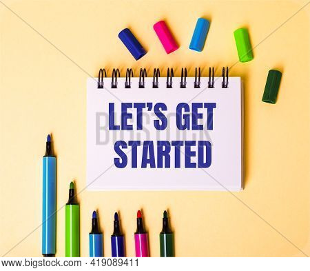 The Words Let Is Get Started Written In A White Notebook On A Beige Background Near Multi-colored Ma