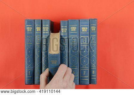 Books Of Classical Literature On A Red Background. A Man's Hand Extracts A Book From The Collected W