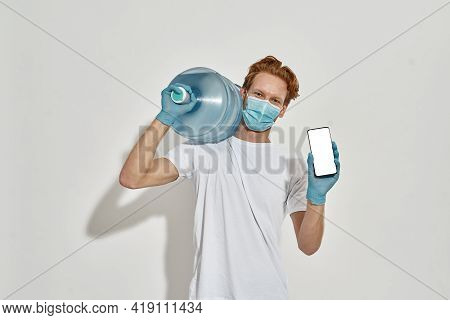 Picture Of A Young Slim Deliveryman Carrying A Huge Bottle Of Water On His Shoulder, Holding A Smart