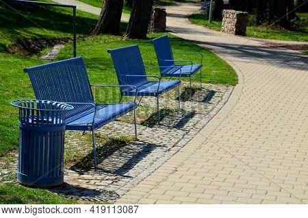 Park Benches In Light Blue Made Of Metal Strips Similar To A Lattice. Trash Can And Beautiful Lawn W